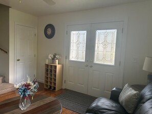 A living room with a wooden coffee table, a black leather sofa, a decorative vase with flowers, a small shelf with a plant, and a double door with frosted glass panels.
