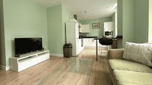 A modern living room with a flat-screen TV, a glass coffee table, a sofa, and a kitchen area with white cabinets and appliances. - Beautiful 2 bed Flat in City Centre (Sheffield)