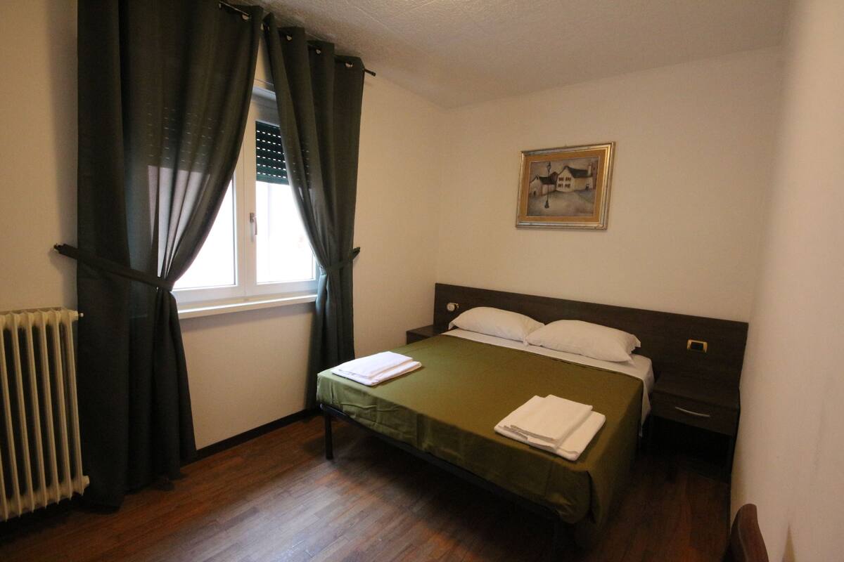 A bedroom with a bed, a window with curtains, a radiator, a nightstand, and a painting on the wall.