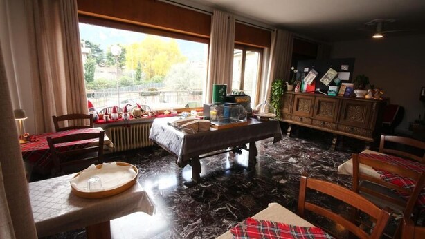 A room with a dining table, chairs, a radiator, a window with curtains, and a cabinet with various items. - R&B Casa Wanda since 1999 (Riva del Garda)