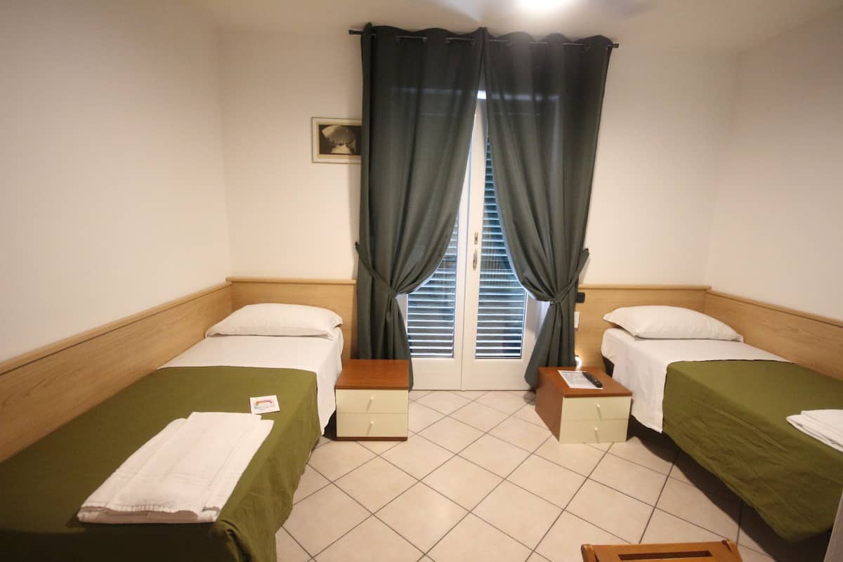 A room with two single beds, a wooden headboard, a small bedside table with a lamp, a window with curtains, and a door with a handle.