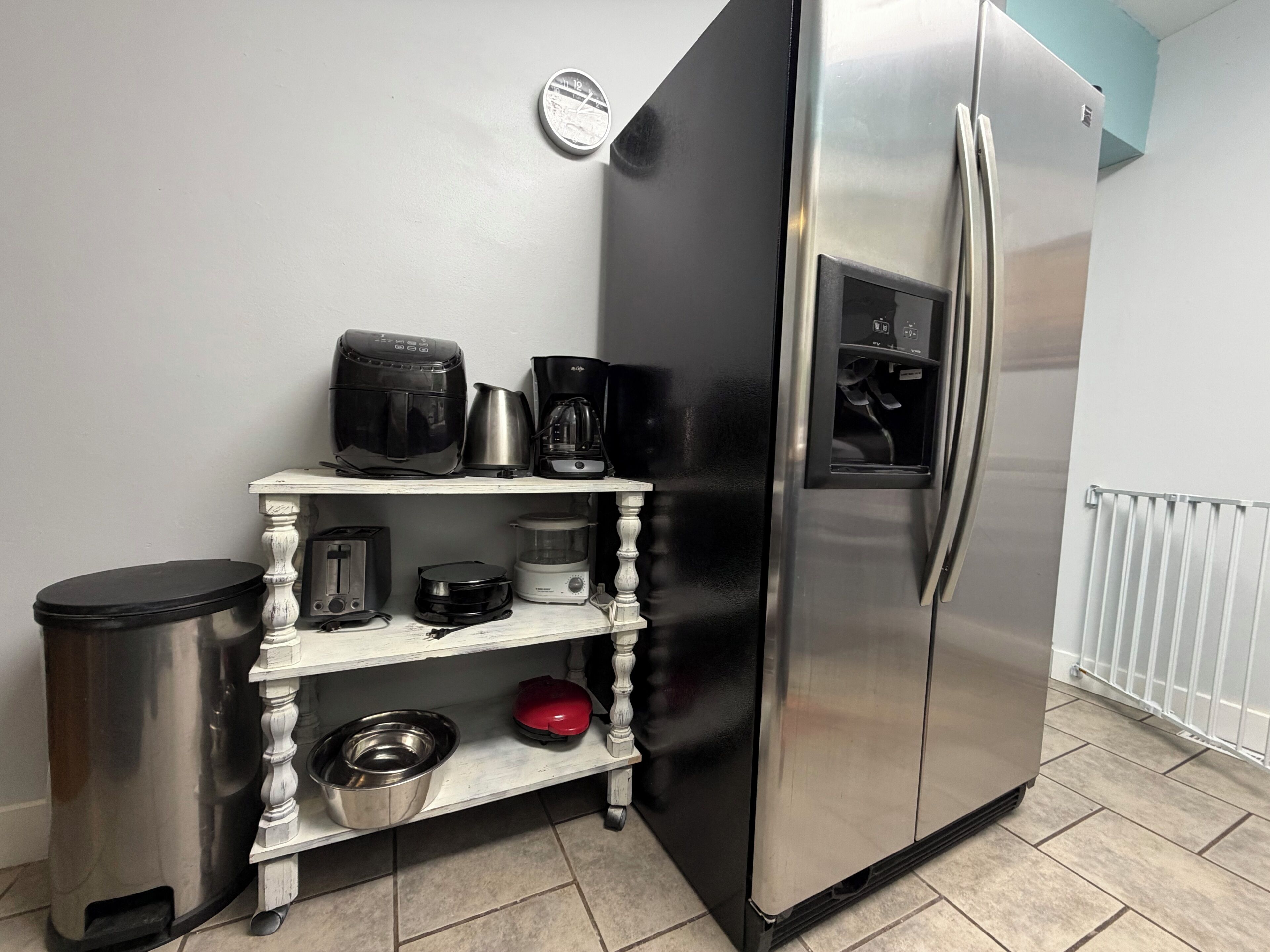 A kitchen with a stainless steel refrigerator, a coffee maker, a kettle, a toaster, and a pet bowl on a white shelf.