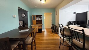 Un comedor con mesa y sillas de madera, una cocina con refrigerador y cafetera, y una estantería con libros y objetos decorativos.