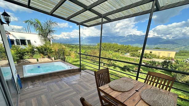 Una terraza de madera con un jacuzzi, una mesa con sillas y vistas a montañas y vegetación.