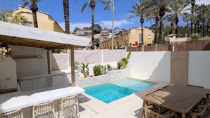 Un área junto a la piscina con mesa y sillas de madera, un espacio de descanso cubierto y una piscina azul cristalina.