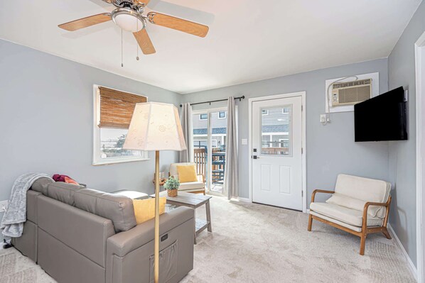 A living room with a ceiling fan, a sofa, a chair, a lamp, a television, and a door leading to a balcony. - Bayview Condo Short Walk to Beach (Ocean City)