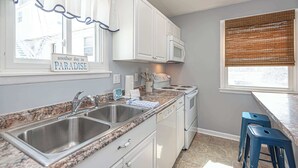 Een keuken met witte kastjes, een granieten aanrecht, roestvrijstalen spoelbakken, een magnetron en een fornuis. Er staan twee blauwe barkrukken en er is een raam met jaloezieën.