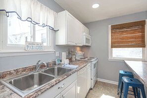 A kitchen with white cabinets, a granite countertop, stainless steel sinks, a microwave, and a stove. There are two blue bar stools and a window with blinds. - Bayview Condo Short Walk to Beach (Ocean City)