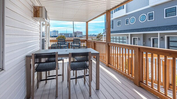 Een overdekt terras buiten met een tafel en stoelen, een houten balustrade en uitzicht op gebouwen achtergrond.