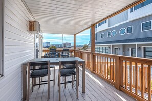 A covered outdoor deck with a table and chairs, a wooden railing, and a view of buildings in the background. - Bayview Condo Short Walk to Beach (Ocean City)