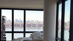 A modern living room with a large window, a stack of books, and a view of a city skyline across a body of water. - 2-bedroom apartment in Greenpoint Brooklyn with stunning views (Brooklyn)
