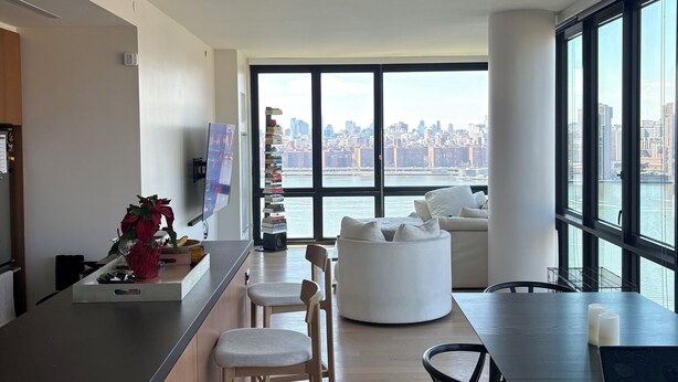 A modern living space with a kitchen area, dining table, and seating. A large window offers a view of a cityscape and water. - 2-bedroom apartment in Greenpoint Brooklyn with stunning views (Brooklyn)