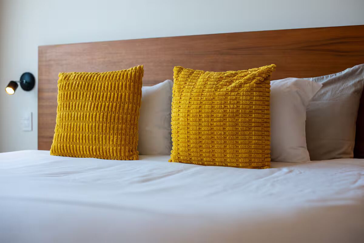 Una cama con ropa de cama blanca, dos almohadas amarillas con textura y un cabecero de madera.