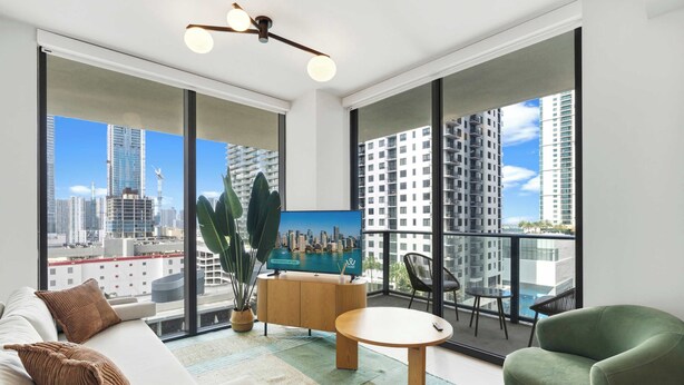 A modern living room with a sofa, a coffee table, a TV, and a balcony with a view of the city. - 2BR/2BA | Pool  |  8 Min to Port of Miami Cruises (Miami)