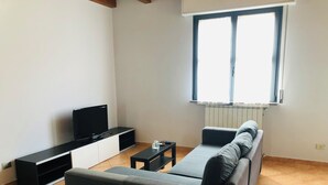 Ein Wohnzimmer mit einer Couch, einem Fernseher auf einem Schrank, einem kleinen Tisch und einem Fenster mit Jalousien.