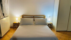 Ein ordentlich bezogenes Bett mit Holz-Kopfteil, zwei Nachttische mit Lampen und ein Heizkörper links.