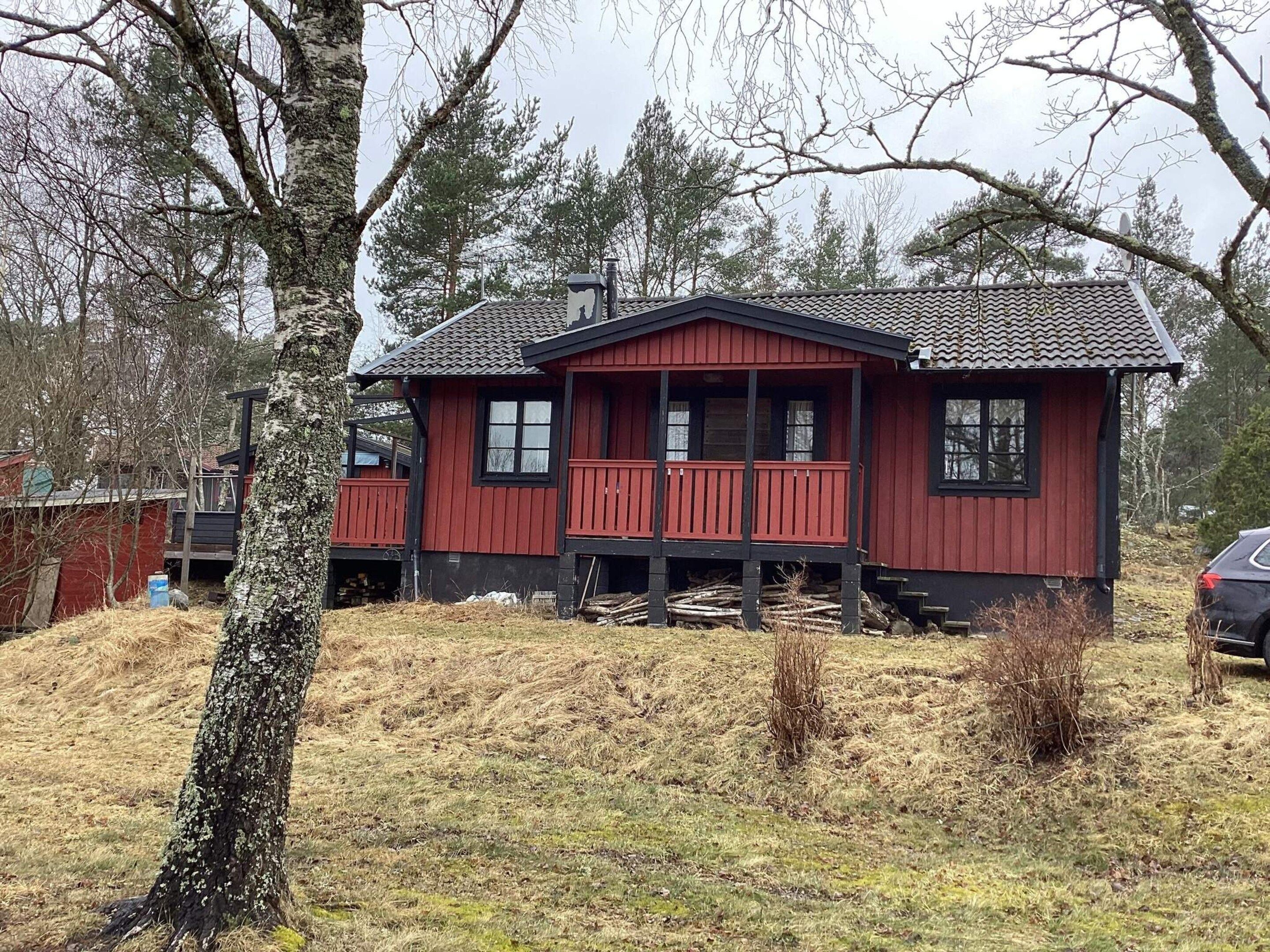 Una cabaña roja de madera con un porche, rodeada de árboles y un área verde.