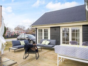 A covered outdoor patio with a wooden deck, two grey sofas, a black chair, and a table with a patterned tablecloth. - 4 Star Holiday Home in Korsor (Korsor)