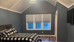 Ein Schlafzimmer mit einem Bett, einem Schreibtisch, einem Fenster mit Jalousien, einer Chandelüster und einer Holzbalkendecke.