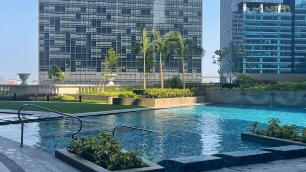 Un bâtiment moderne doté d’une façade en verre, d’un espace piscine avec une terrasse en bois et de palmiers.