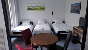 A hotel room with two single beds, a round wooden table, a red chair, and a TV on a wooden shelf. - Quadruple hotel room (Lully)