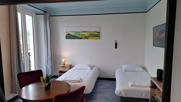 A hotel room with two beds, a dining table, a small plant, and a painting on the wall. - Quadruple hotel room (Lully)