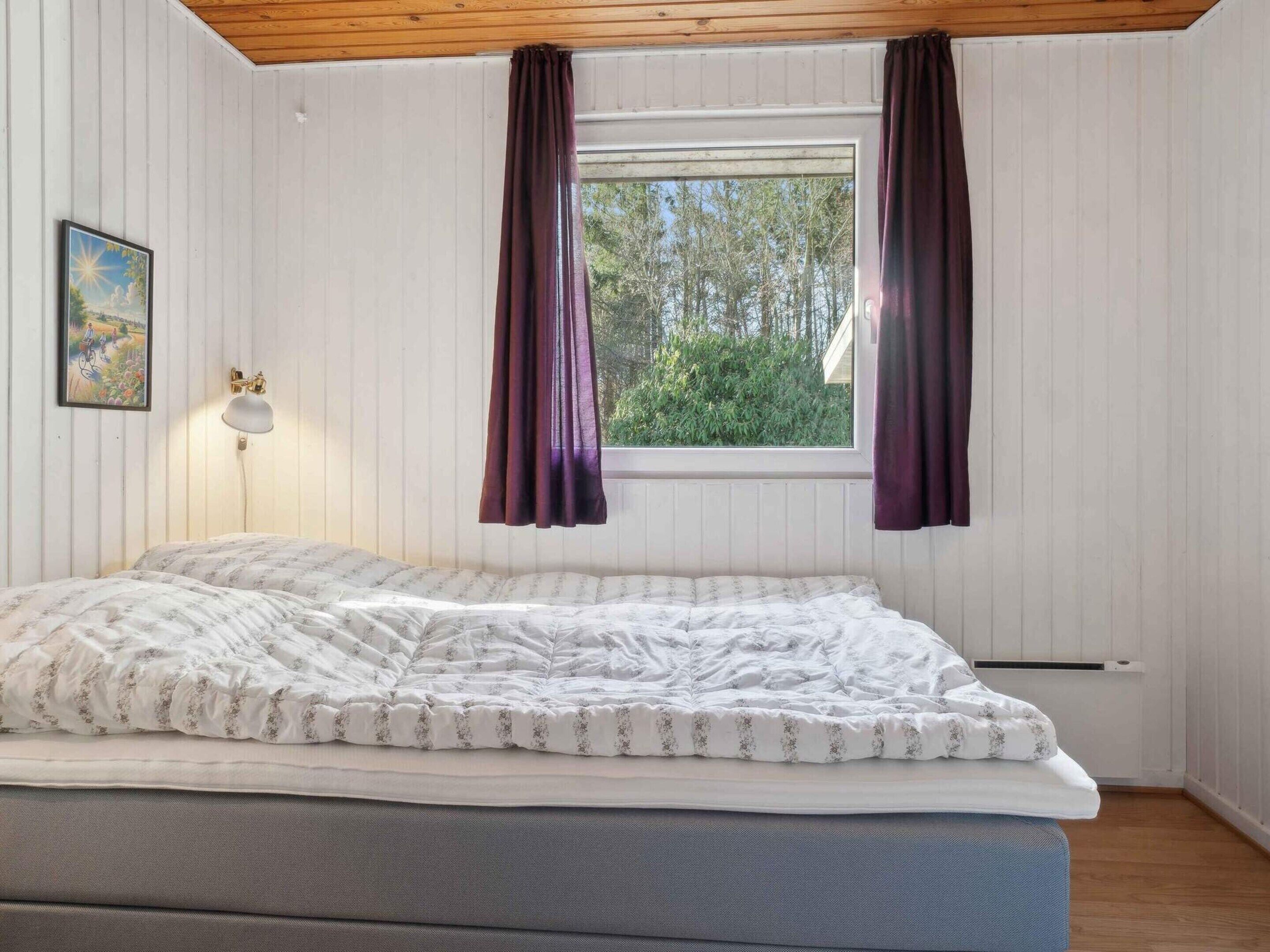 Une pièce avec un lit, une fenêtre avec des rideaux, une lampe fixée au mur et un tableau.