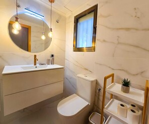 A bathroom with a white marble wall, a round mirror, a white sink, a toilet, and a small shelf with a plant. - Unique by iulia (Oropesa del Mar)