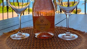 Eine Flasche Sandara Sekt auf einem Tisch mit zwei Gläsern, vor einer Küstenlandschaft.