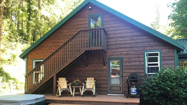 A wooden cabin with a staircase leading to a deck, two chairs, a grill, and a hot tub. - Cozy cabin in the woods with internet, hot tub,fire pit, grille, gas stove (Walton)