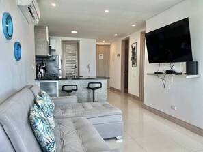 Ein modernes Wohnzimmer mit einer grauen Eckcouch, einem flachbildfernseher an der Wand und einer Küchenzeile im Hintergrund.