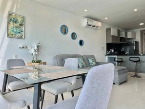 Ein modernes Wohnzimmer mit einem Glastisch, grauen Sofas und einer Küchenzeile mit Edelstahlgeräten.