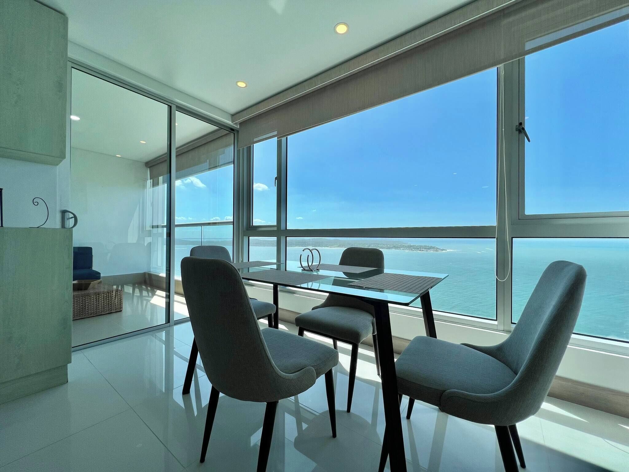 A modern room with a glass wall, a table with chairs, and a view of the sea.