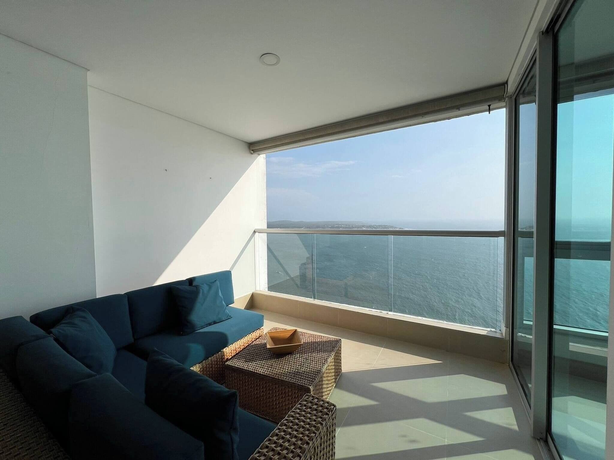 A balcony with a sofa and wicker furniture, overlooking a body of water.