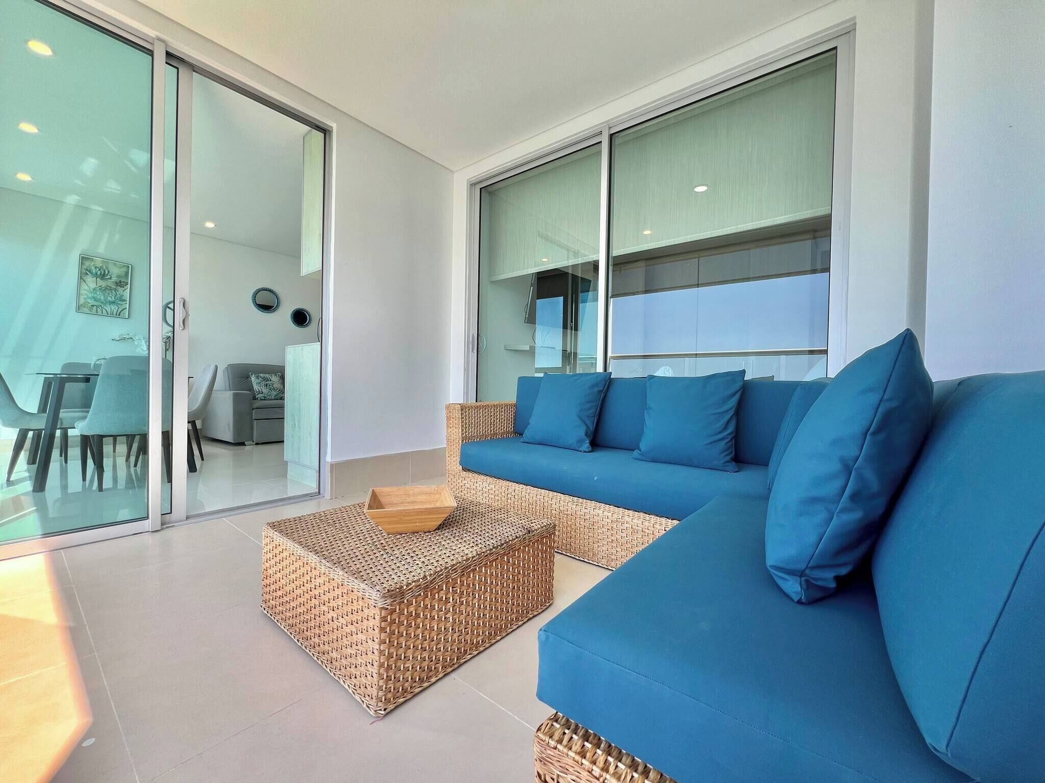 A modern living room with a blue sofa, wicker ottoman, and glass sliding doors leading to a dining area.