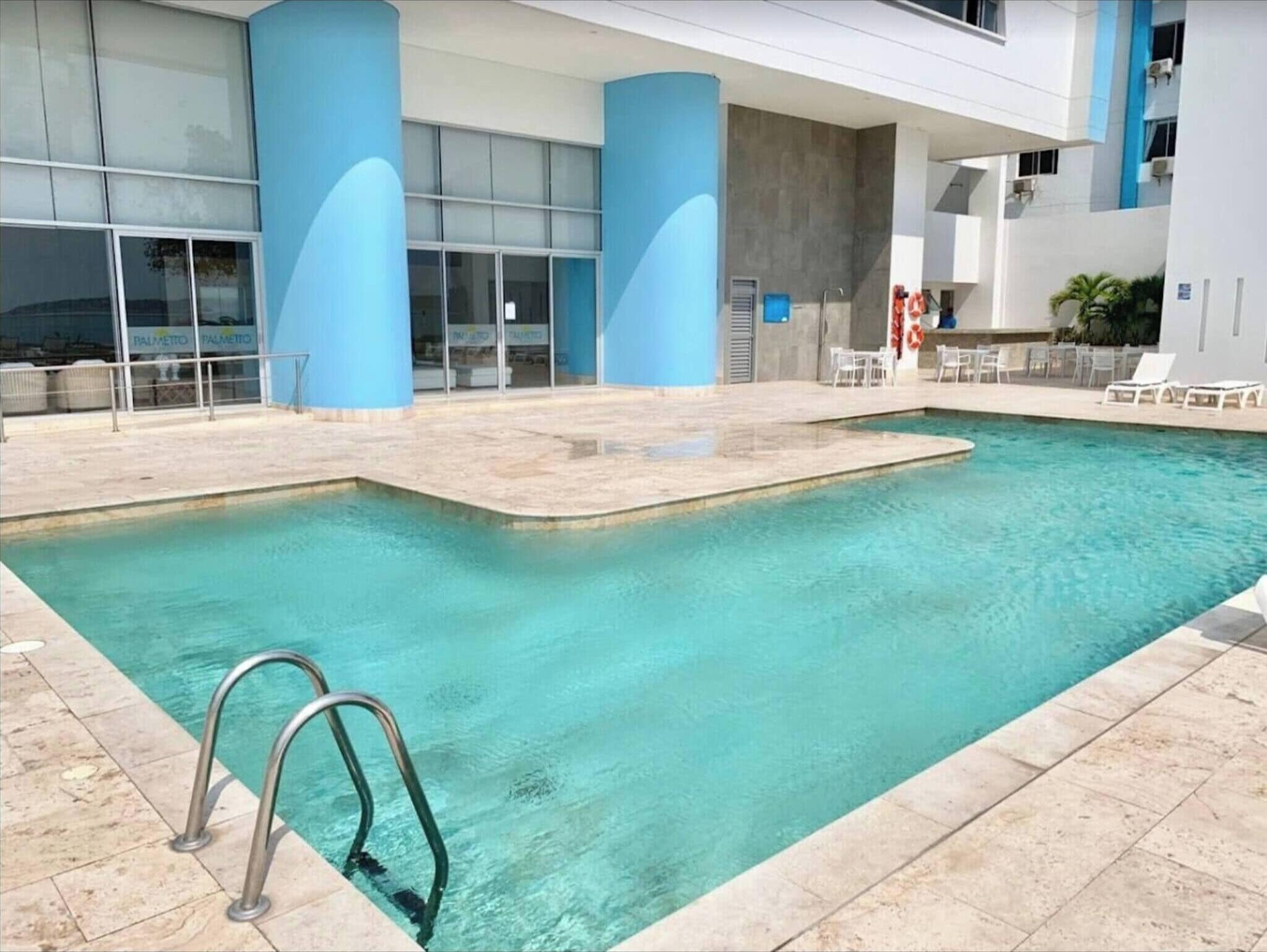 Uma piscina com uma escada de metal, rodeada por um deck revestido em azulejos e um edifício com grandes colunas azuis e janelas de vidro.
