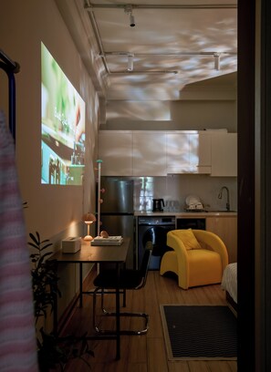 Eine kompakte Küche mit Kühlschrank, Mikrowelle und Kaffeemaschine. Es gibt eine kleine Essecke mit Tisch und Stühlen, sowie einen gelben Hocker.
