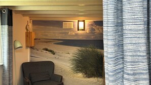 Una habitación con sofá a cuadros, una lámpara, vista a un paisaje desértico y una cortina a rayas.
