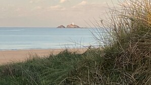 Un paisaje costero con un primer plano de hierba, una playa arenosa y un faro a lo lejos.