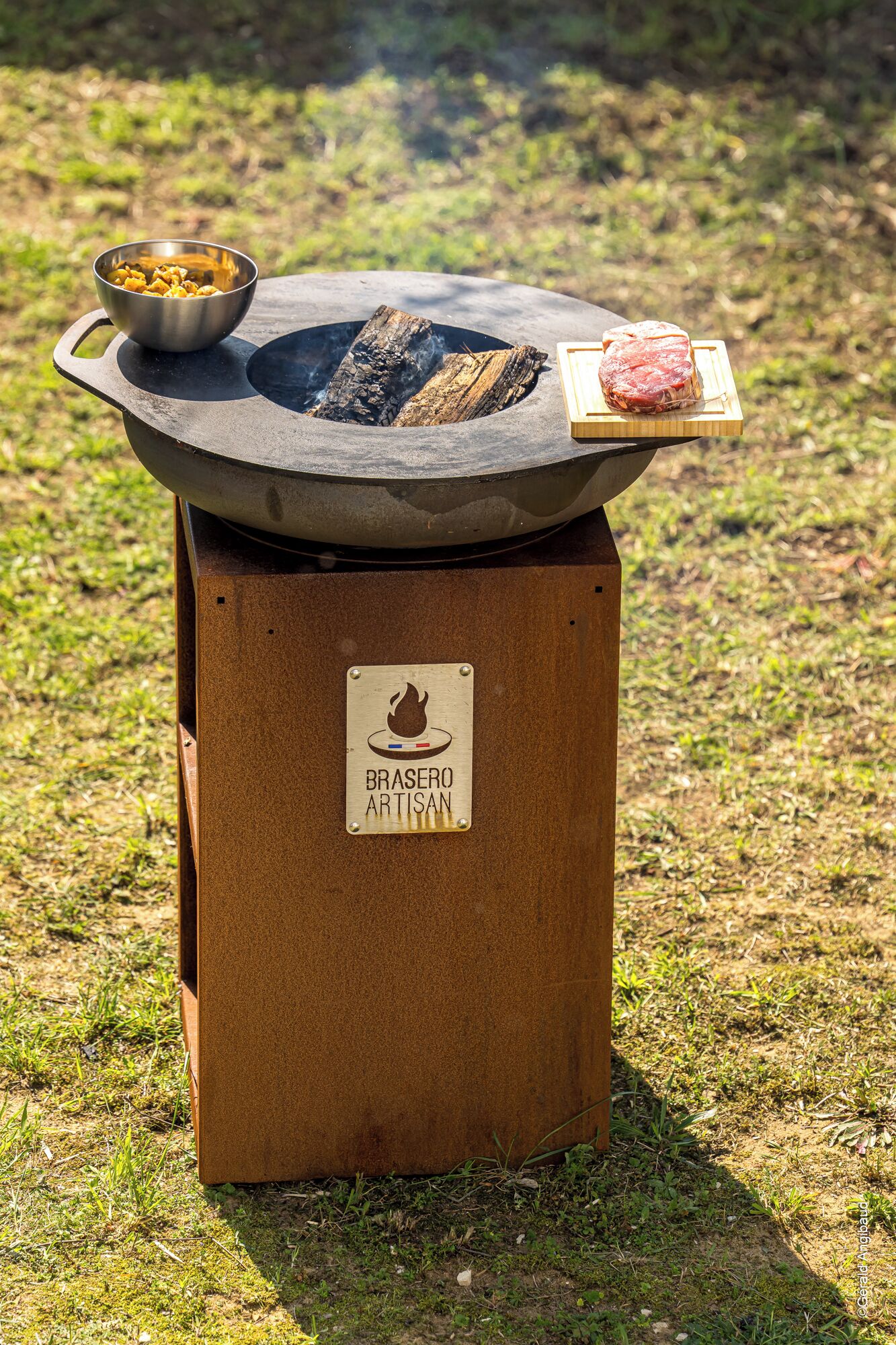 Ein tragbarer Grill mit einer Metallschale und einer Holztischplatte mit Fleisch, beschriftet 'BRASERO ARTISAN'.
