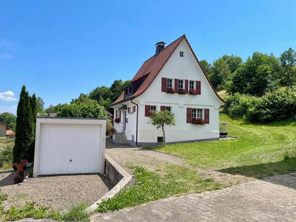 Une maison à deux étages avec un toit recouvert de tuiles rouges, une façade blanche et des volets rouges. On y trouve un garage fermé et un petit arbre devant.
