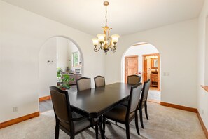 A dining room with a dark wooden table and chairs, a chandelier, and a view through an archway to another room.