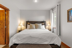 Quarto com cabeceira de madeira, roupa de cama branca, duas luminárias de apoio e janela com cortinas.