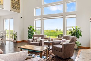 A spacious living room with large windows, a wooden coffee table, two armchairs, and a small side table.