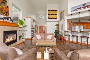 A spacious living room with a fireplace, comfortable seating, and a kitchen area with wooden cabinets and bar stools. - Bridger Mountain Vista-Private, Clean, Hot Tub, Stunning Rocky Mountain Views (Bozeman)