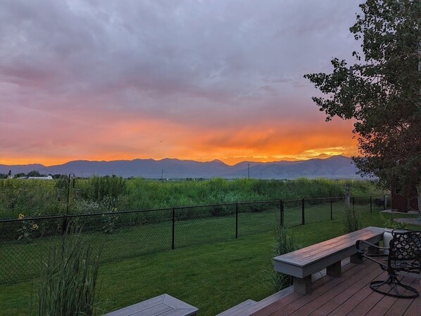A patio with a wooden bench and a table, overlooking a grassy area with a fence and mountains in the background.