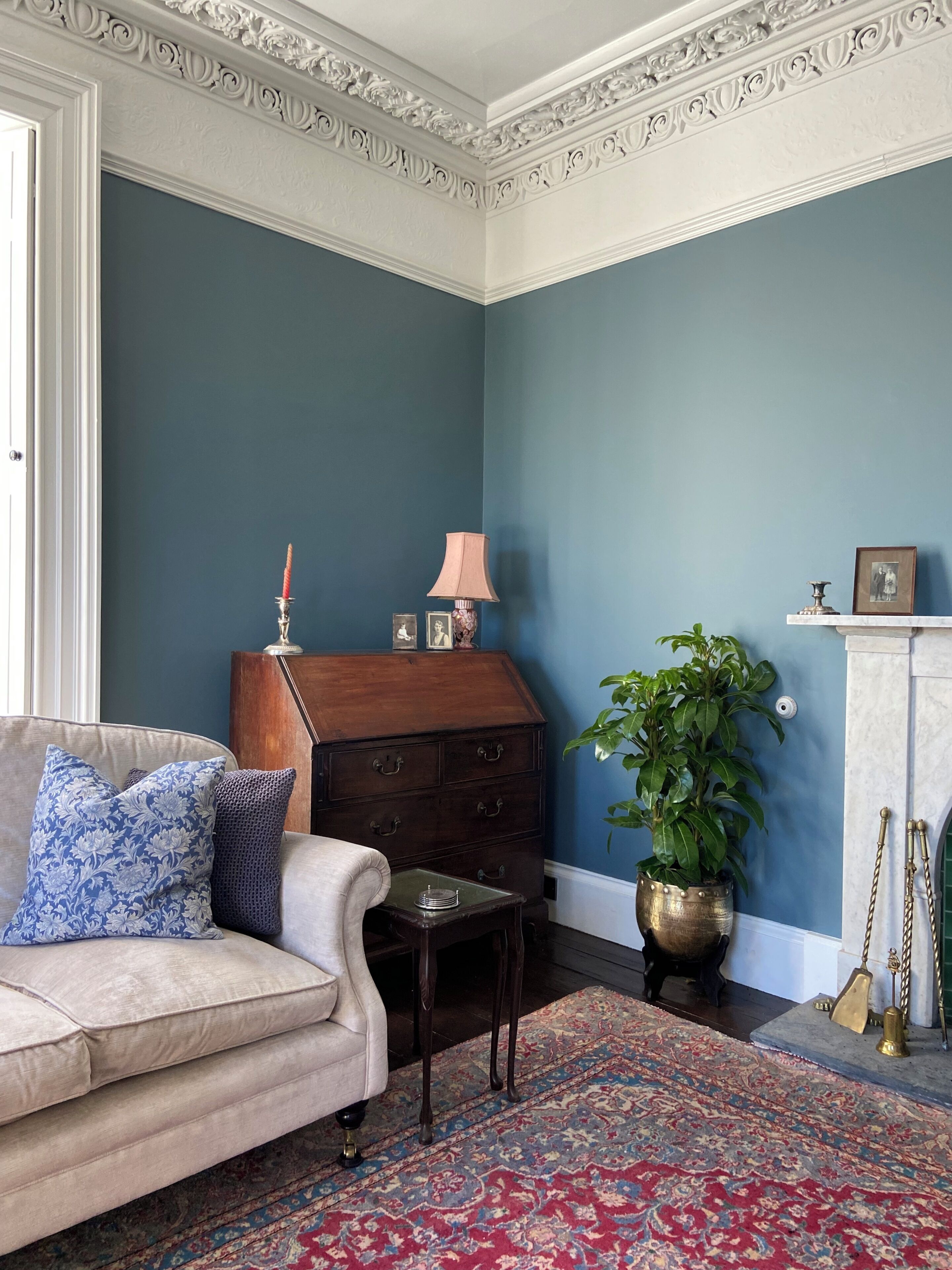 A room with a blue wall, a wooden chest of drawers, a white sofa with patterned cushions, a small table, a potted plant, and a patterned rug.