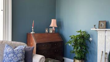 A room with a blue wall, a wooden chest of drawers, a white sofa with patterned cushions, a small table, a potted plant, and a patterned rug.