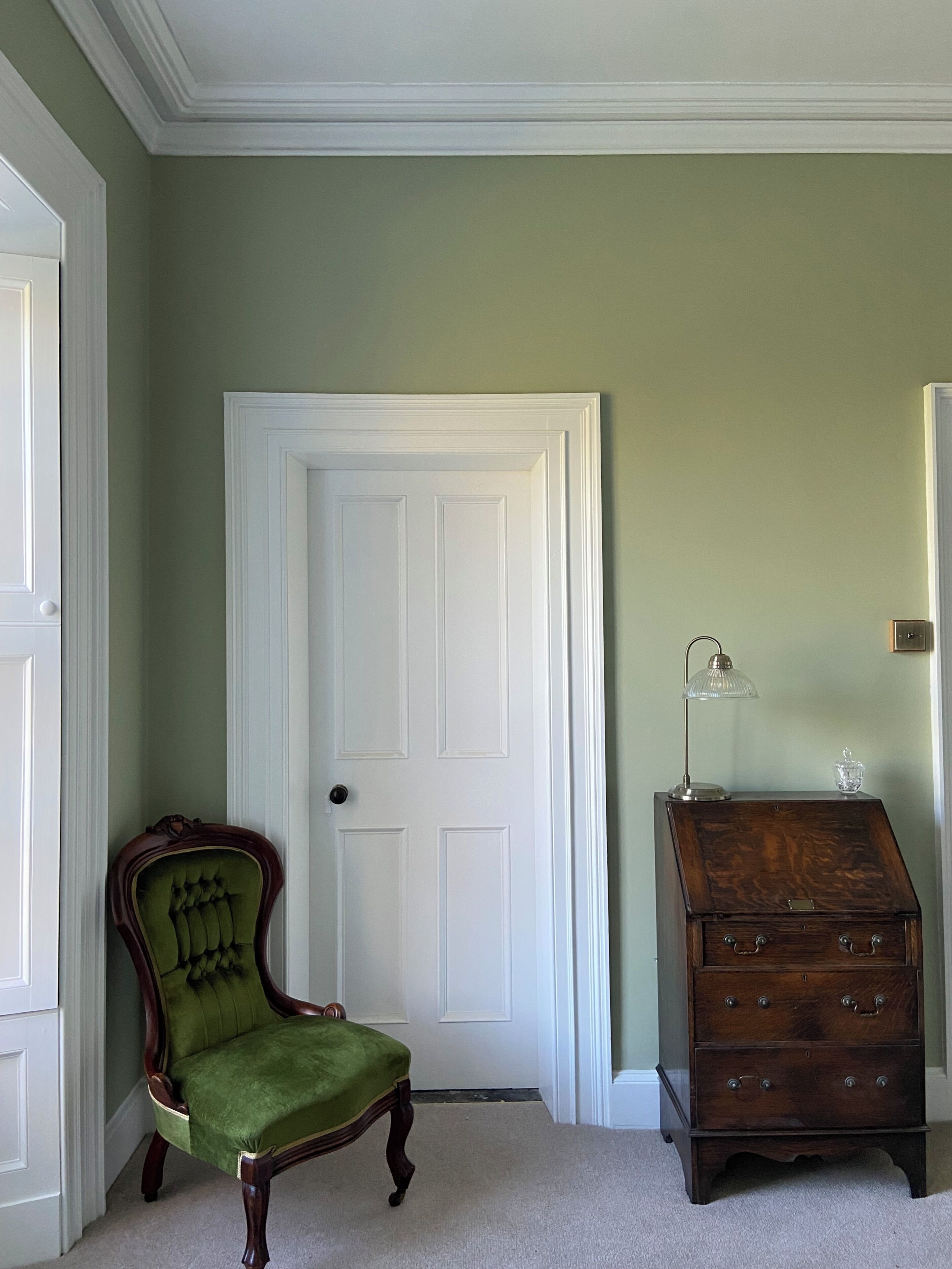 Una habitación con pared verde, puerta blanca, sillón tapizado en verde y cómoda de madera con cajones.
