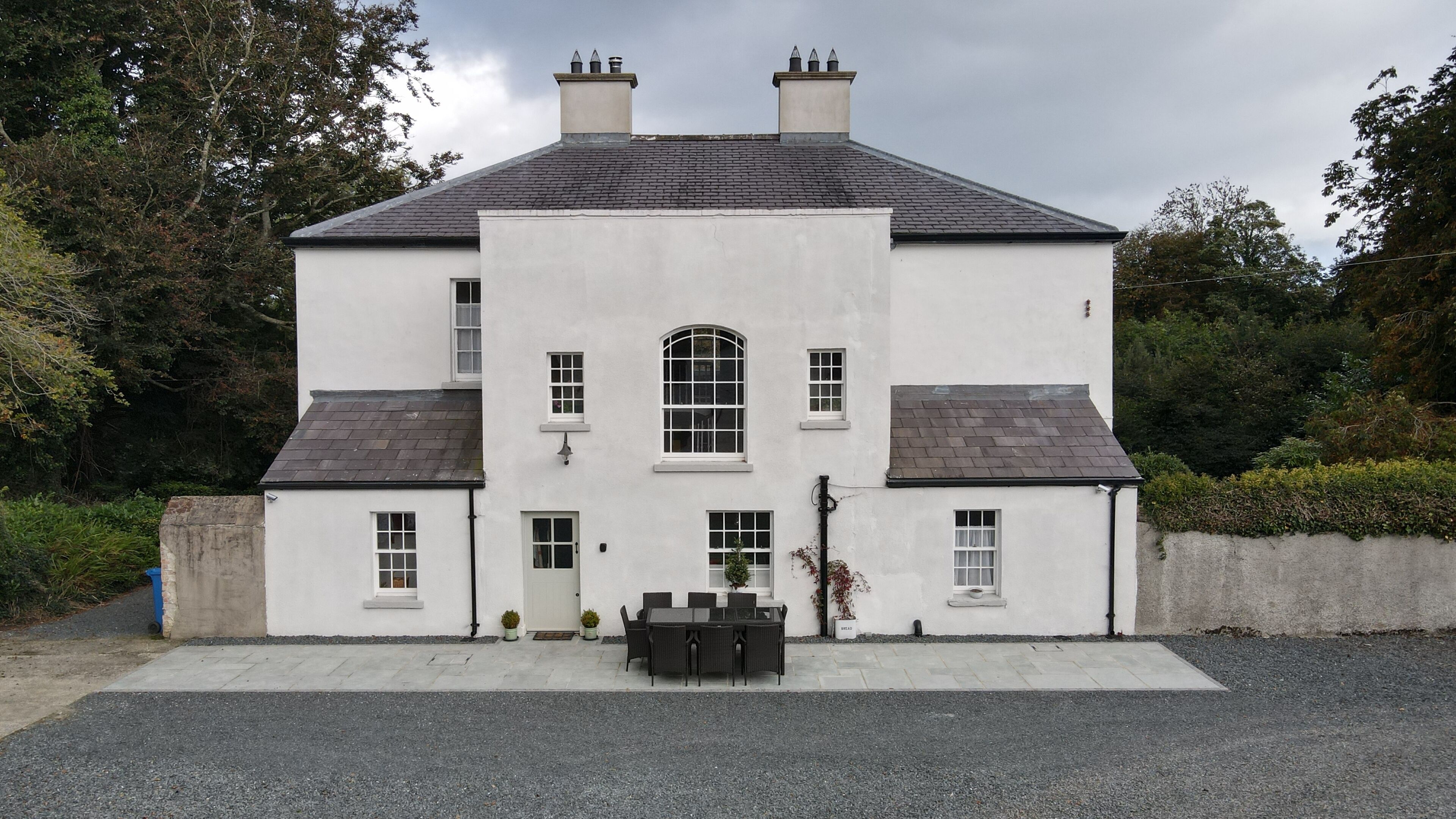 A two-story building with a symmetrical facade, a central arched window, and a front porch with a table and chairs.